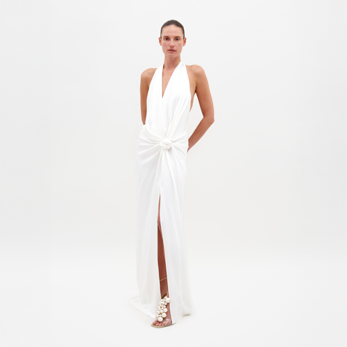 Person standing against a white background, wearing the Torgiano Dress White, which features a sleeveless halter design with a knot detail at the waist and a front slit. Pre-order now to ensure your dress is shipped by February 15th, 2025.