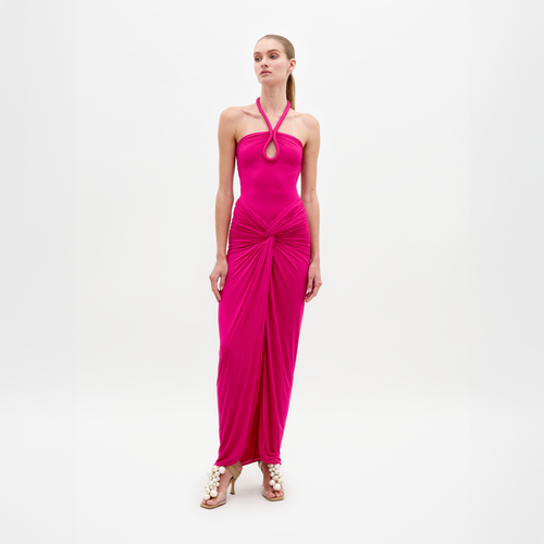 A woman stands in the Tori Dress Fuchsia, featuring a bright pink halter neck design with ruched detailing, paired with statement beige heels on a plain white background. This stylish ensemble is available for pre-order until February 15th, 2025.