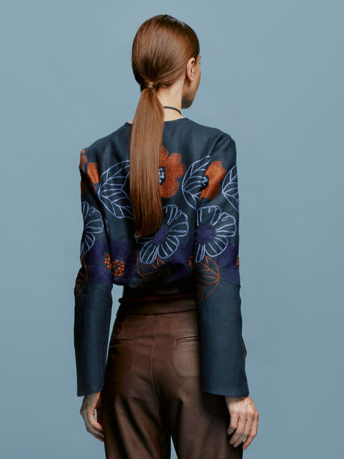 A person stands against a blue background, adorned in the Tosca Blouse Navy Ochre Flower Embroidery with its elegant keyhole neckline, complemented by brown pants and a belt featuring keys and leather accessories. This stylish ensemble could easily be the highlight of fashion week on September 2nd or any day in 2024.