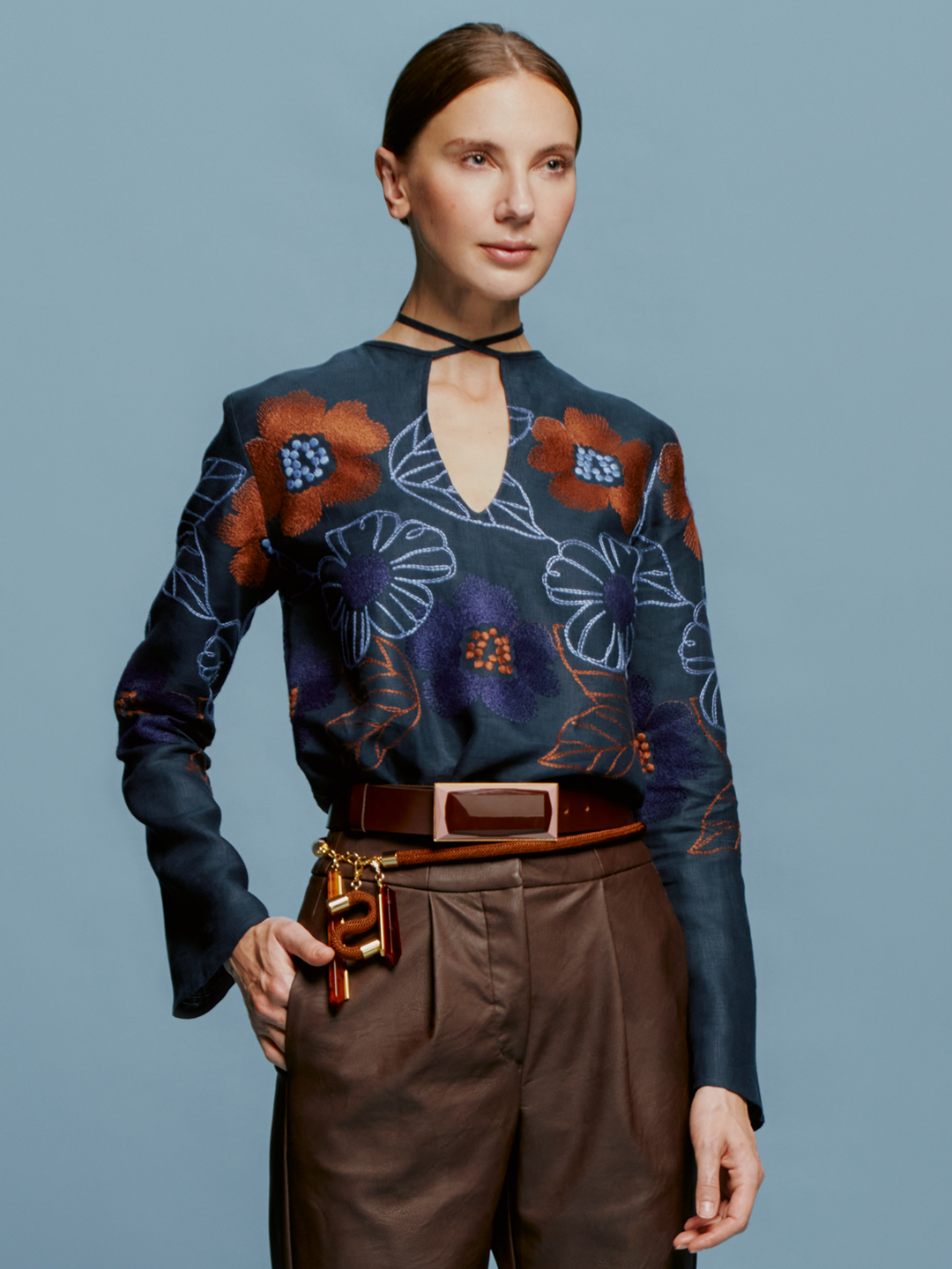 A person stands against a blue background, adorned in the Tosca Blouse Navy Ochre Flower Embroidery with its elegant keyhole neckline, complemented by brown pants and a belt featuring keys and leather accessories. This stylish ensemble could easily be the highlight of fashion week on September 2nd or any day in 2024.