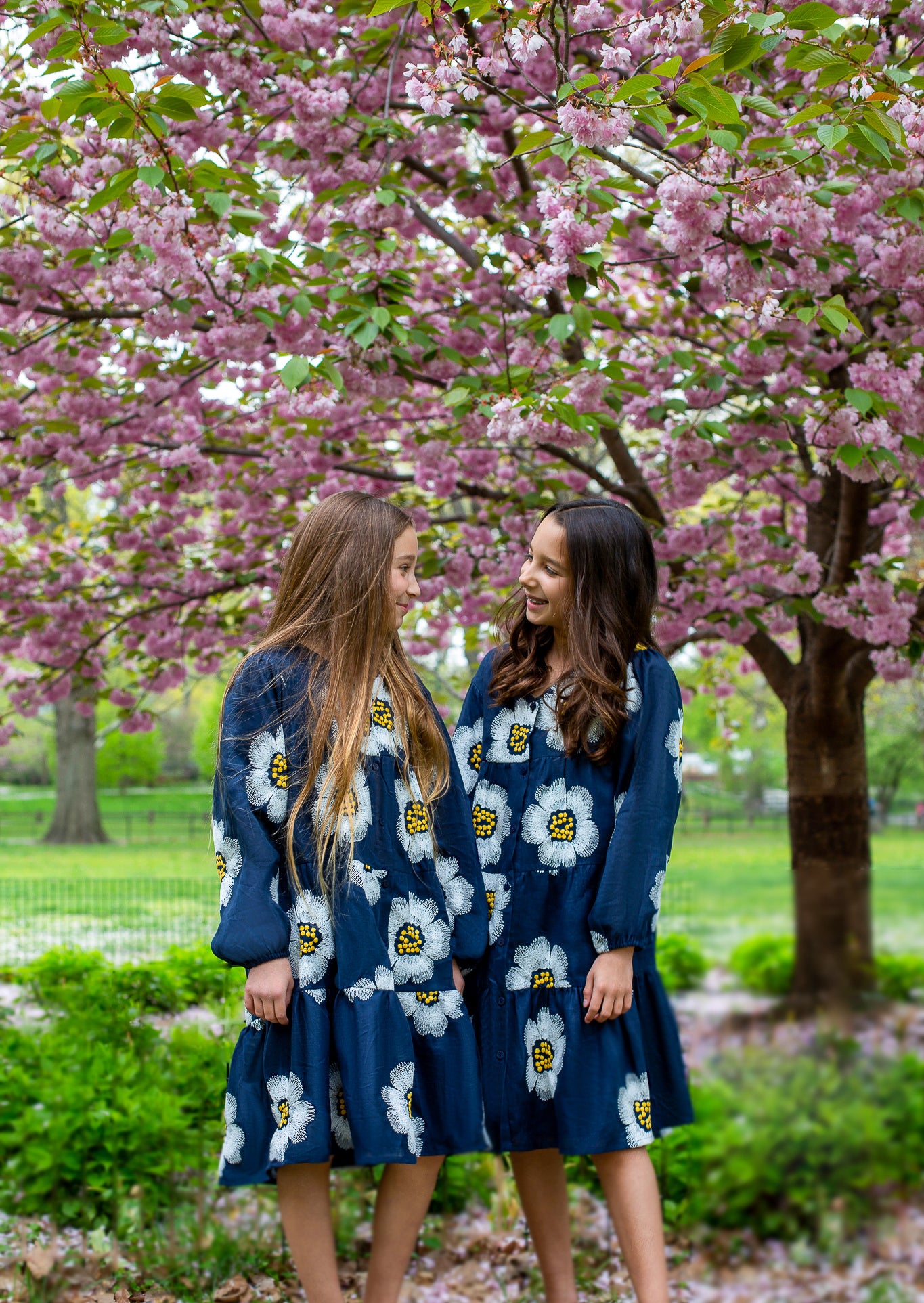 Two girls in matching blue floral dresses stand under a blooming cherry blossom tree, smiling at each other.