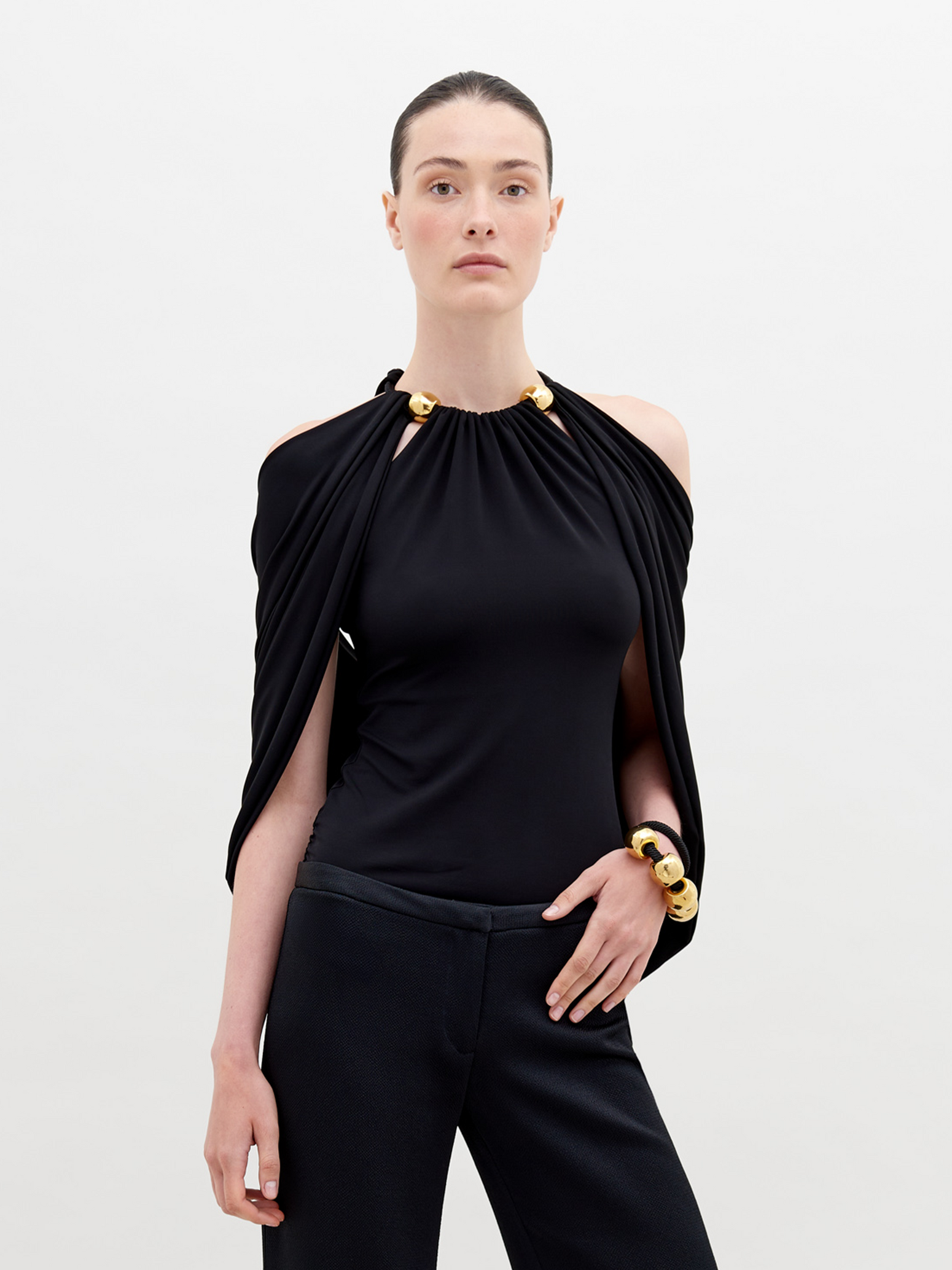 A person with tied-back hair wears a Tabatha Blouse Black with gold accents and a matching gold bracelet, standing against a plain white background. This stylish ensemble is available for pre-order now, with a ship by date of November 15th, 2024.