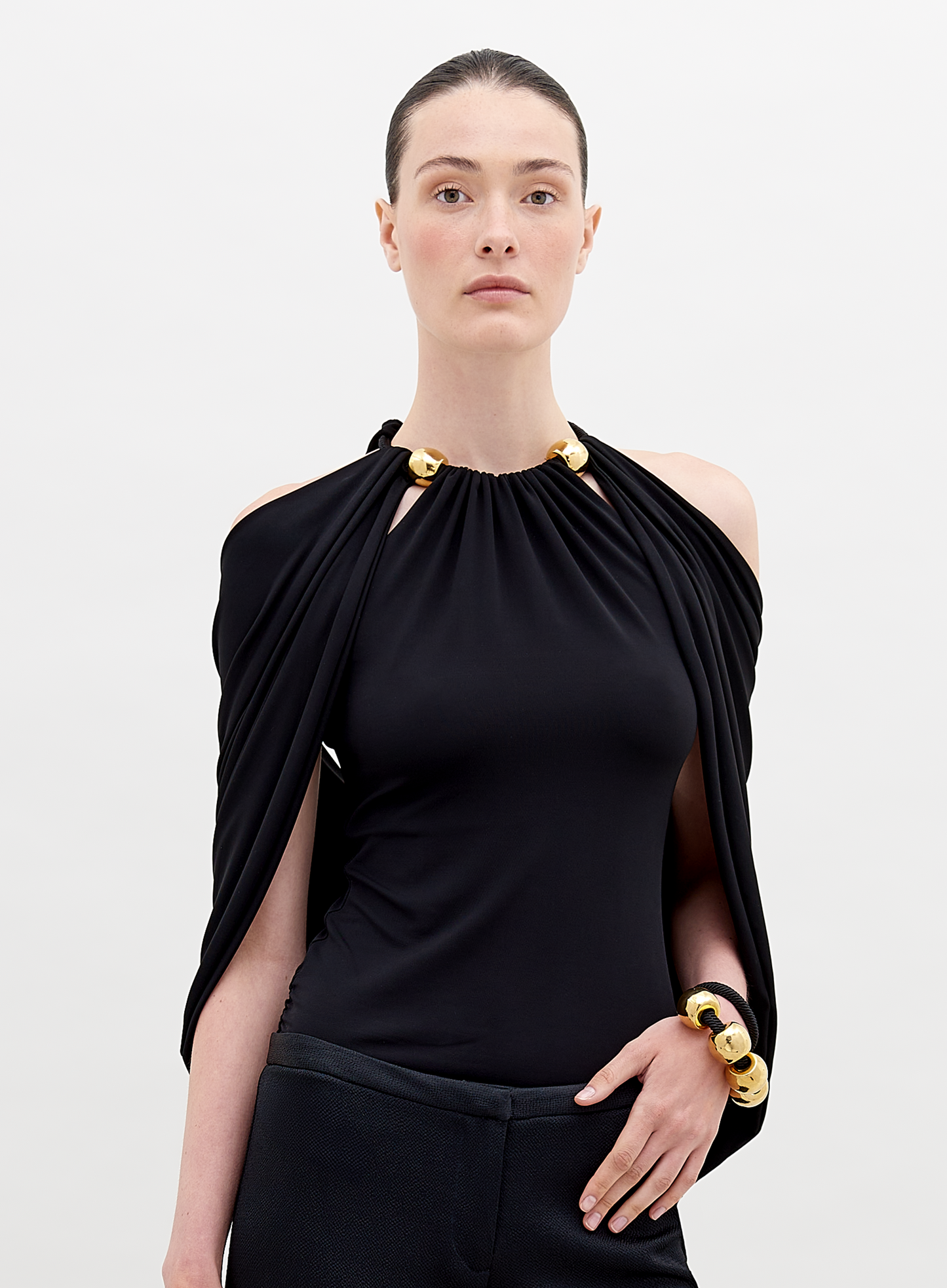 A person with tied-back hair wears a Tabatha Blouse Black with gold accents and a matching gold bracelet, standing against a plain white background. This stylish ensemble is available for pre-order now, with a ship by date of November 15th, 2024.