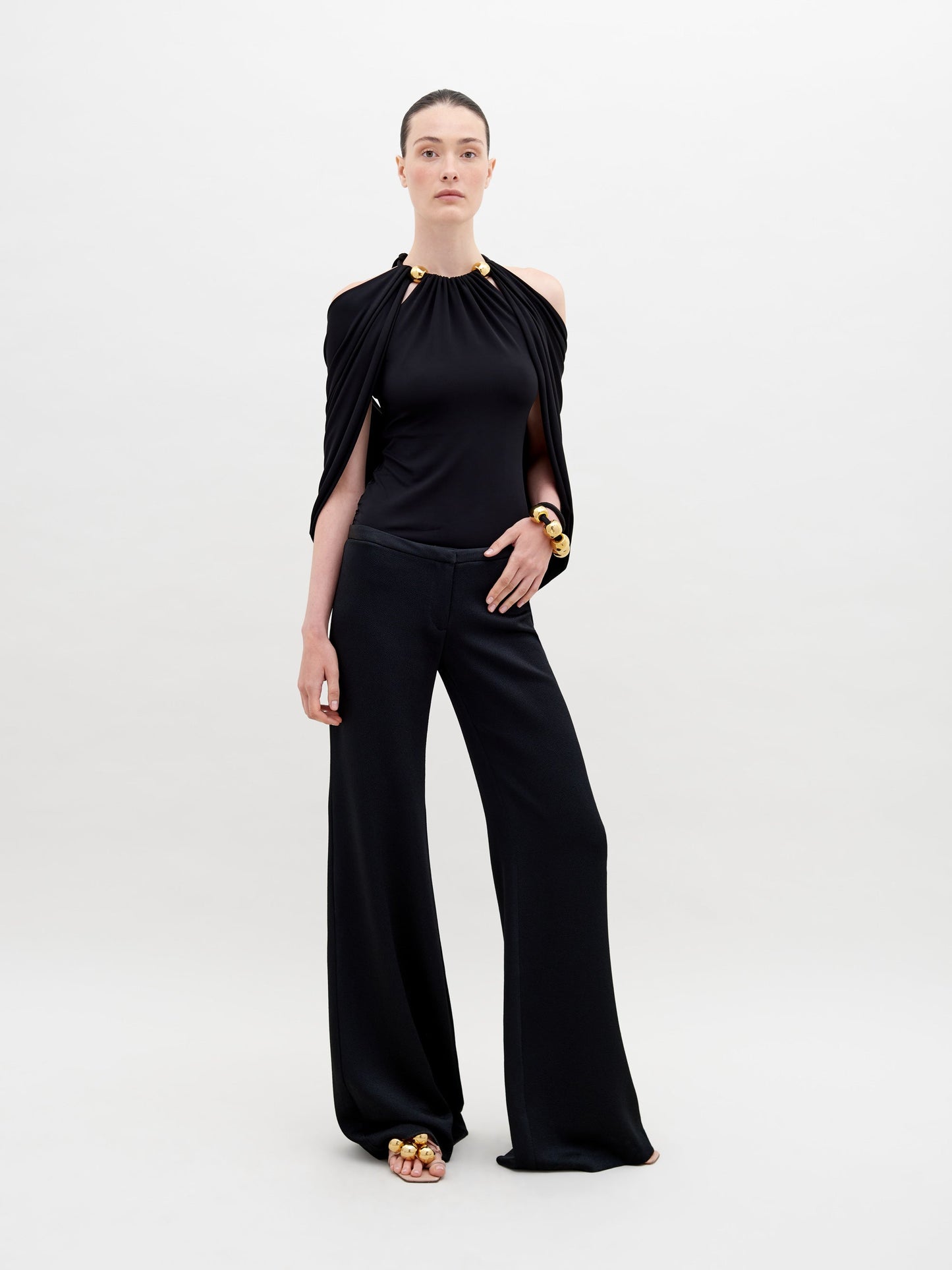 A person stands against a plain background wearing a stylish, sleeveless black top with draped fabric and Ovidia Pant Black, accessorized with gold-toned bracelets and sandals. Available for pre-order now, this ensemble will ship by date in early 2024.