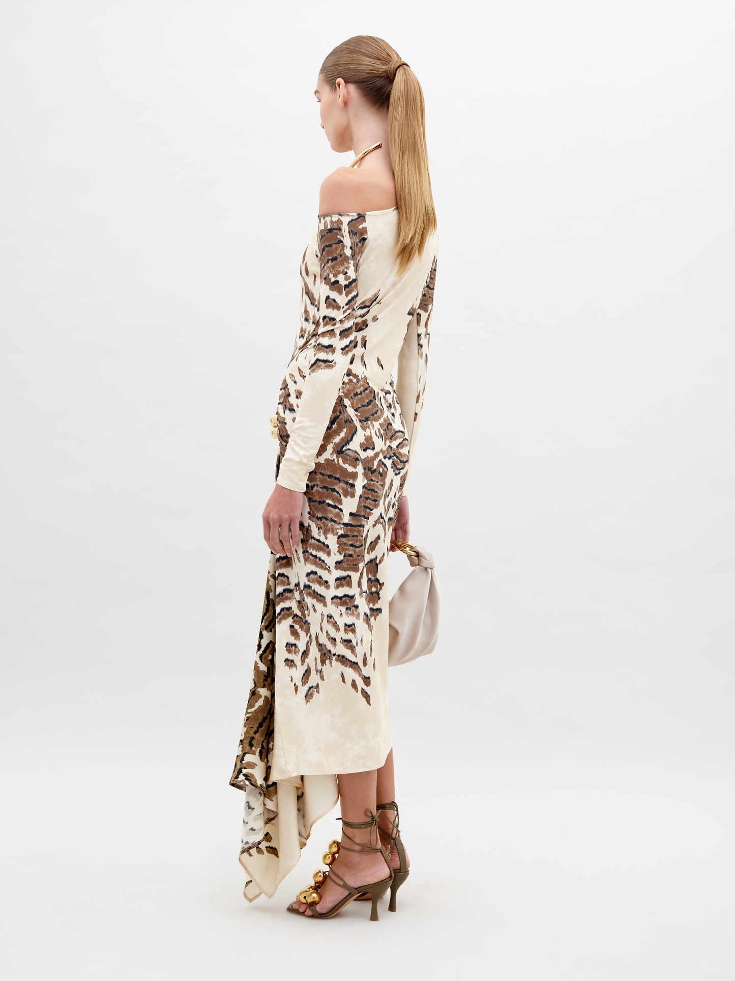 A woman in an off-shoulder, floor-length Tahia Dress Beige Abstract Animal Print stands against a plain background, holding a large beige bag in one hand. She is wearing gold sandals and a gold choker. Mark your calendars for November 15th—this stunning look is available for pre-order ahead of its 2024 launch.