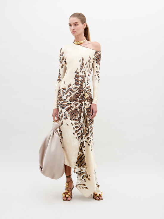 A woman in an off-shoulder, floor-length Tahia Dress Beige Abstract Animal Print stands against a plain background, holding a large beige bag in one hand. She is wearing gold sandals and a gold choker. Mark your calendars for November 15th—this stunning look is available for pre-order ahead of its 2024 launch.