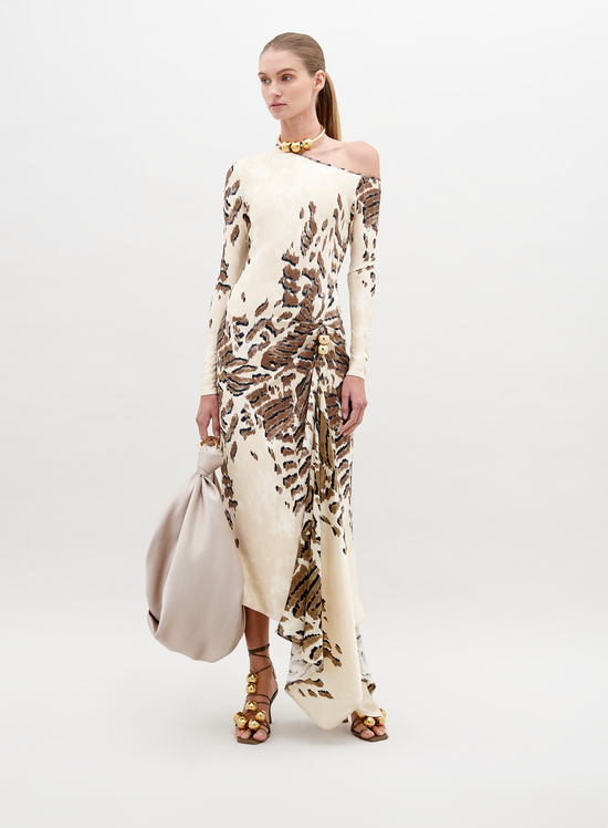 A woman in an off-shoulder, floor-length Tahia Dress Beige Abstract Animal Print stands against a plain background, holding a large beige bag in one hand. She is wearing gold sandals and a gold choker. Mark your calendars for November 15th—this stunning look is available for pre-order ahead of its 2024 launch.