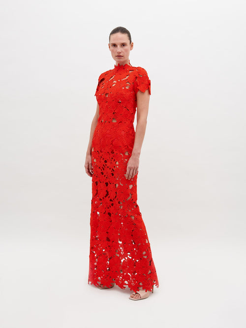 Person wearing a floor-length, red lace Taiana Dress Rouge with a high neckline and short sleeves, standing against a plain white background. Pre-order now to ensure delivery by the November 15th, 2024 ship-by date.