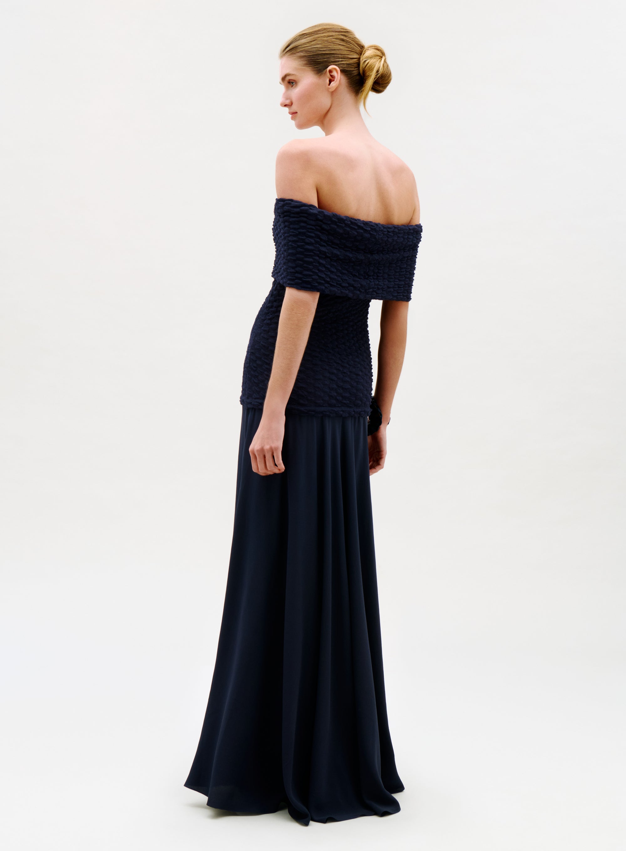 A woman in profile wears the Taylor Dress, featuring an off-the-shoulder, navy blue textured top and a long, flowing skirt. Her hair is in a neat bun against a plain, light background.