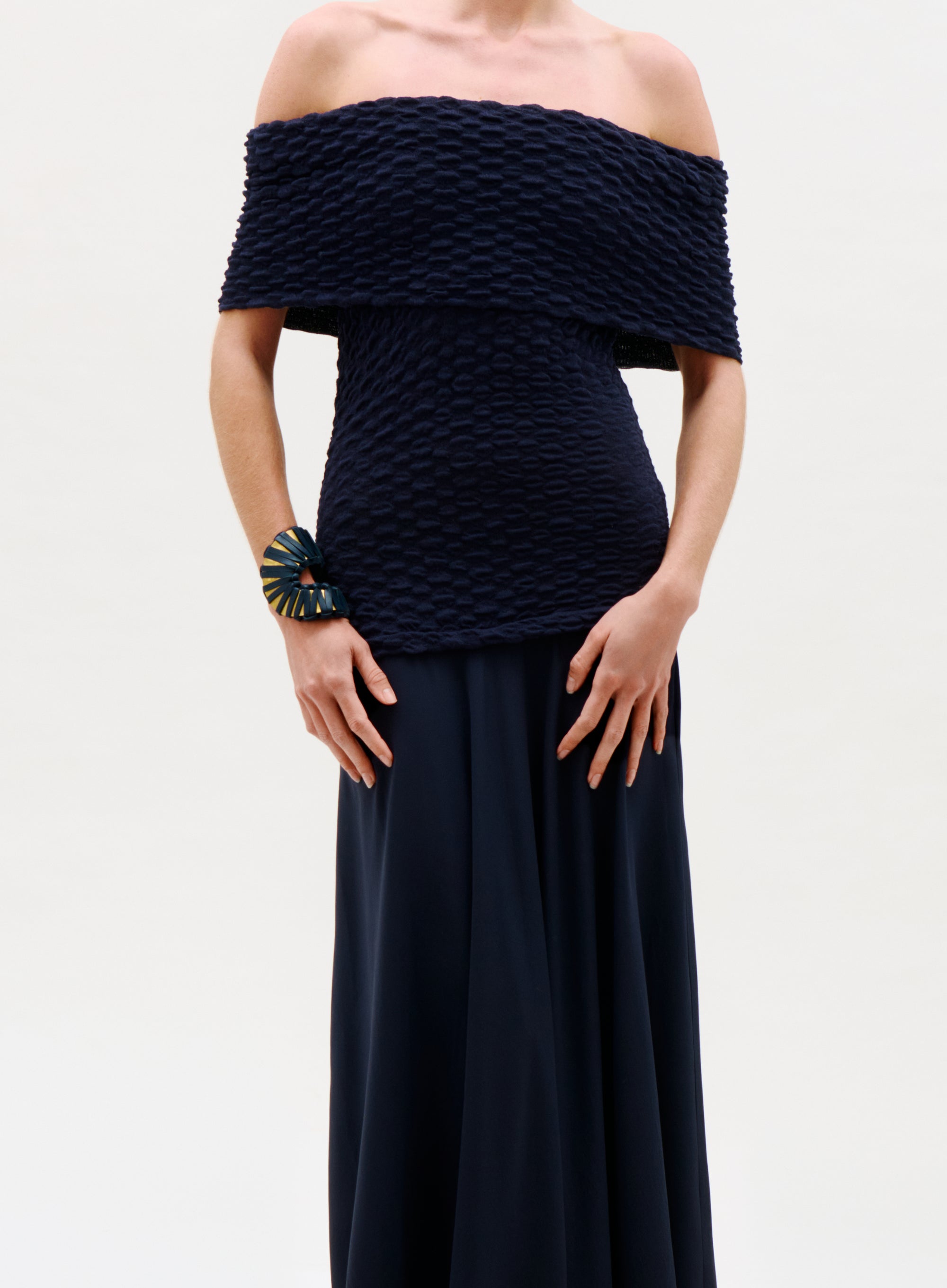 A person wearing the Taylor Dress, an off-the-shoulder textured black outfit paired with a decorative bracelet on their left wrist, stands against a plain light background.