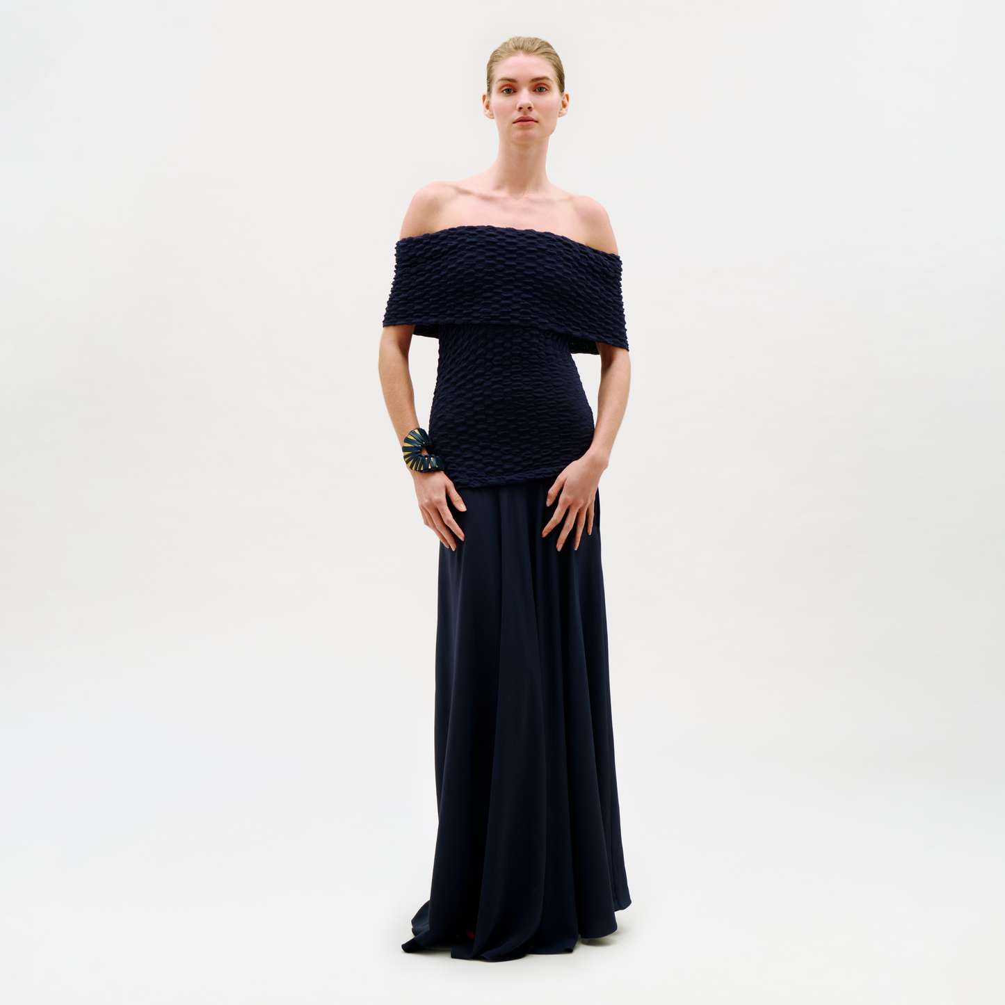 A woman stands against a plain light background, wearing the Taylor Dress—an off-the-shoulder, textured dark dress with a long, flowing skirt. Her hair is pulled back and she accessorizes with a bold statement bracelet on her left wrist.