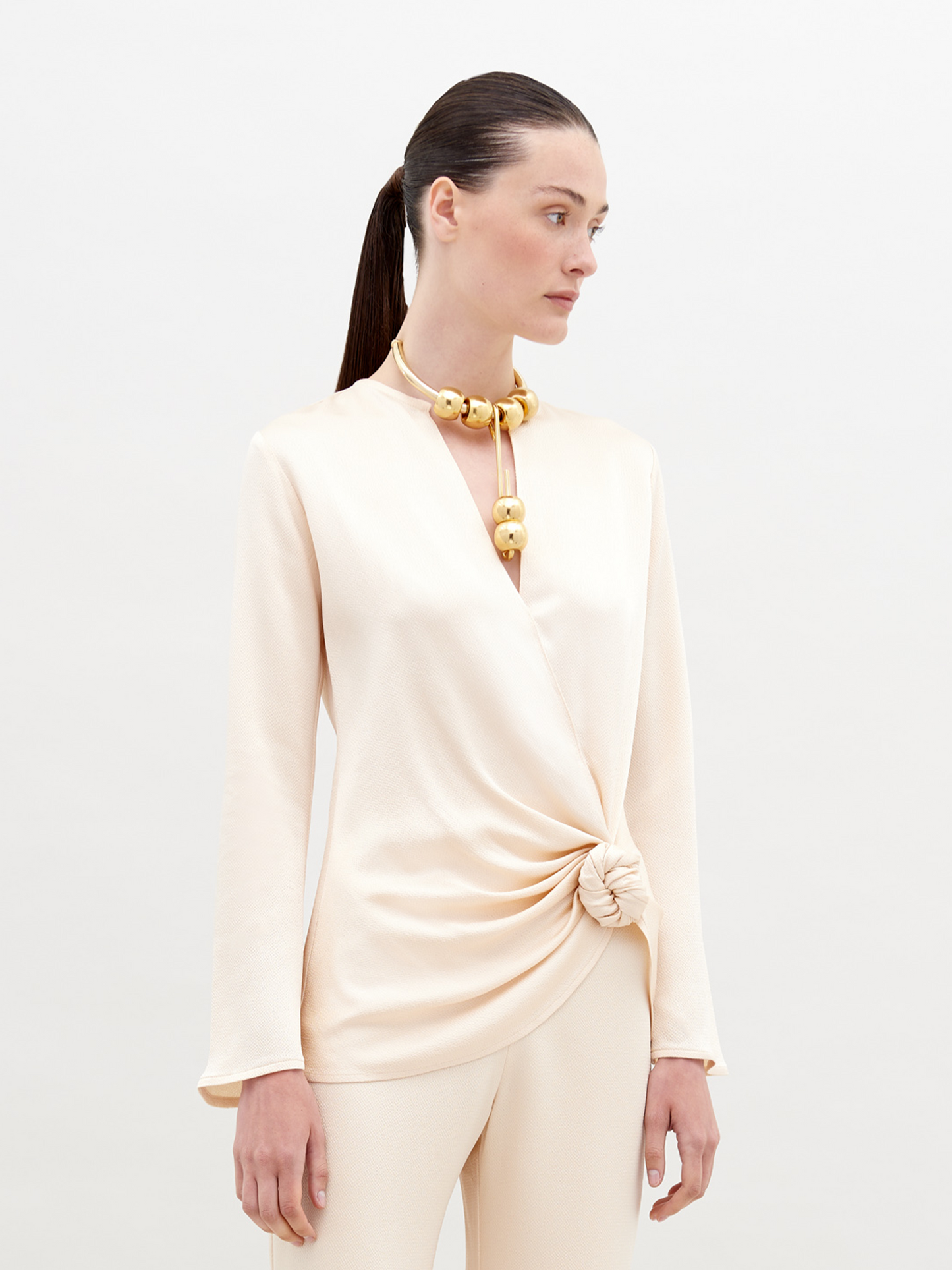 A woman in a cream-colored outfit stands against a plain backdrop. She is wearing a Tayra Blouse Vanilla with a knot detail at the waist and a large statement necklace with round, gold beads. Pre-order now and have it delivered by the ship date of November 15th, 2024.