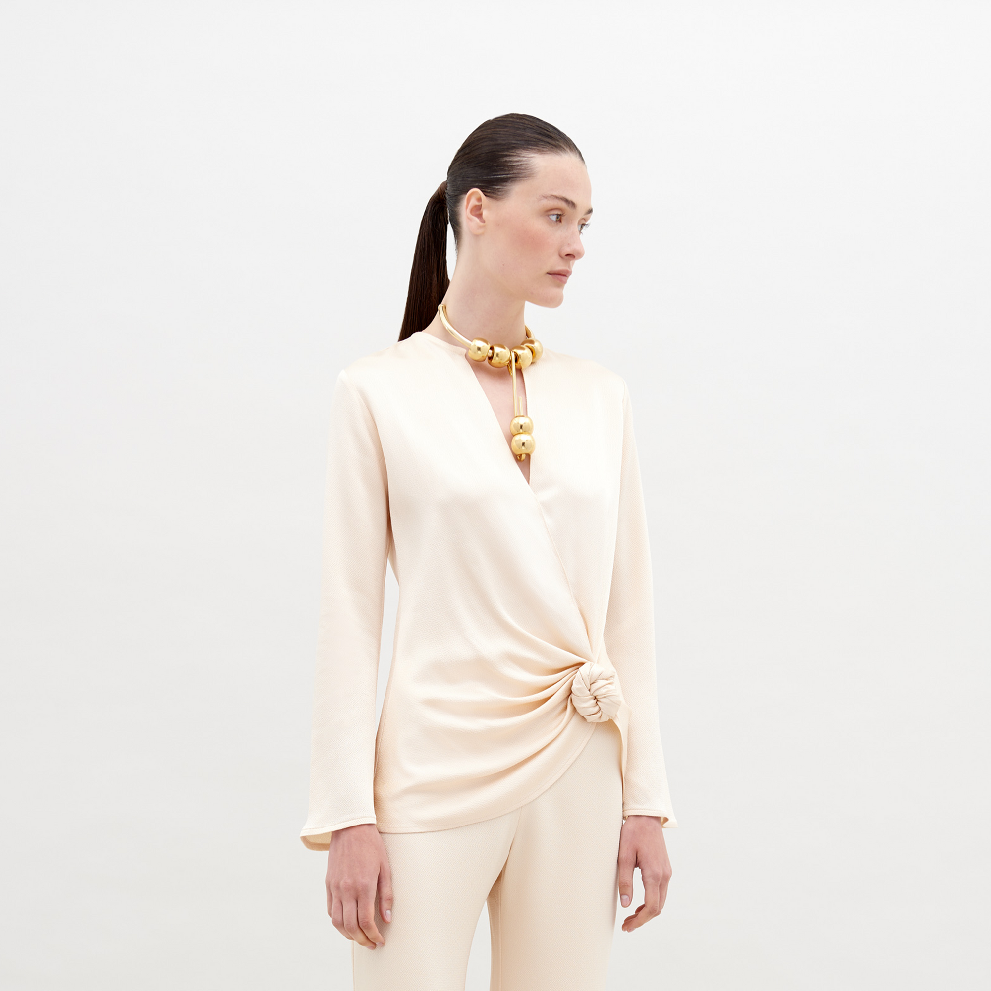 A woman in a cream-colored outfit stands against a plain backdrop. She is wearing a Tayra Blouse Vanilla with a knot detail at the waist and a large statement necklace with round, gold beads. Pre-order now and have it delivered by the ship date of November 15th, 2024.