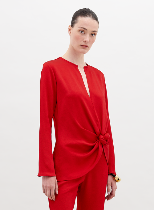 A woman stands in a red outfit with a knot detail on the top. She has her hair pulled back and stares directly into the camera against a plain white background. Available for pre-order now, the Tayra Blouse Rouge will have a ship date of November 15th, 2024.