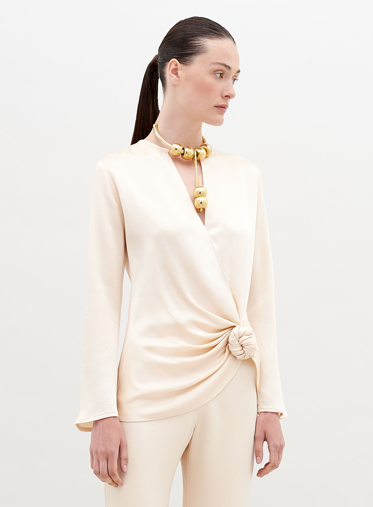 A woman in a cream-colored outfit stands against a plain backdrop. She is wearing a Tayra Blouse Vanilla with a knot detail at the waist and a large statement necklace with round, gold beads. Pre-order now and have it delivered by the ship date of November 15th, 2024.