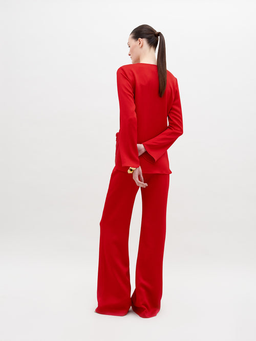 A person stands against a plain background wearing a red long-sleeved top with a knot detail at the waist and matching Palermo Pant Rouge, available for pre-order with a ship by date of November 15th, 2024.