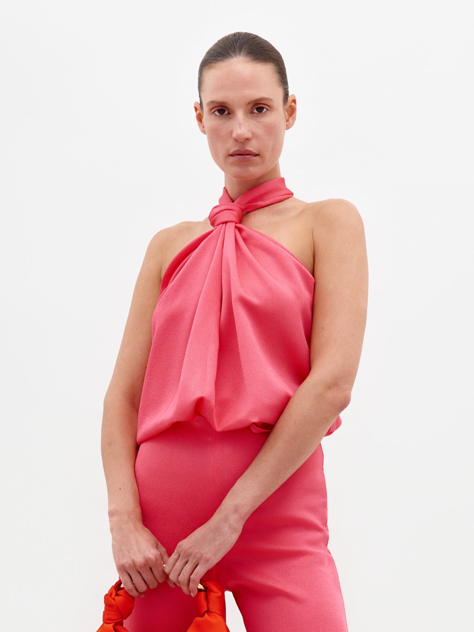 A person stands against a plain background, dressed in the Teagan Blouse Peony—a sleeveless pink top tied at the neck—and matching pink pants, holding a red bag. Available for pre-order now with delivery by February 15th.