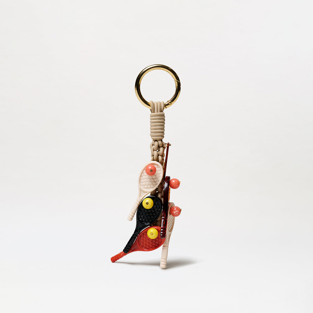 The Tournament Charm is a keychain with a gold ring, beige woven cord, and charms shaped like mini tennis rackets and balls in red, black, and yellow.