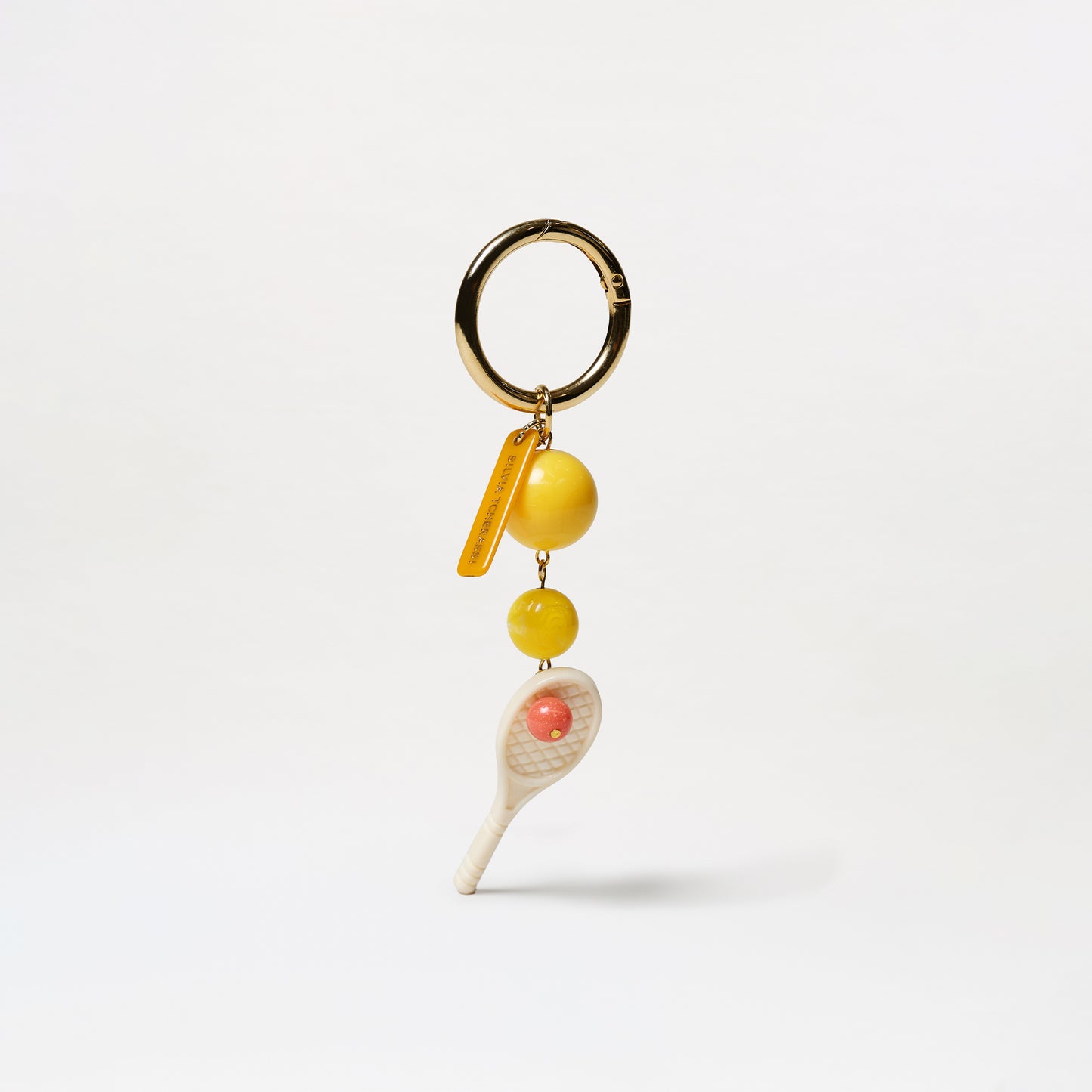 The Deuce Charm keychain features a gold ring, two yellow tennis ball beads, and a white tennis racket charm with a pink ball in the center, all displayed against a plain white background.