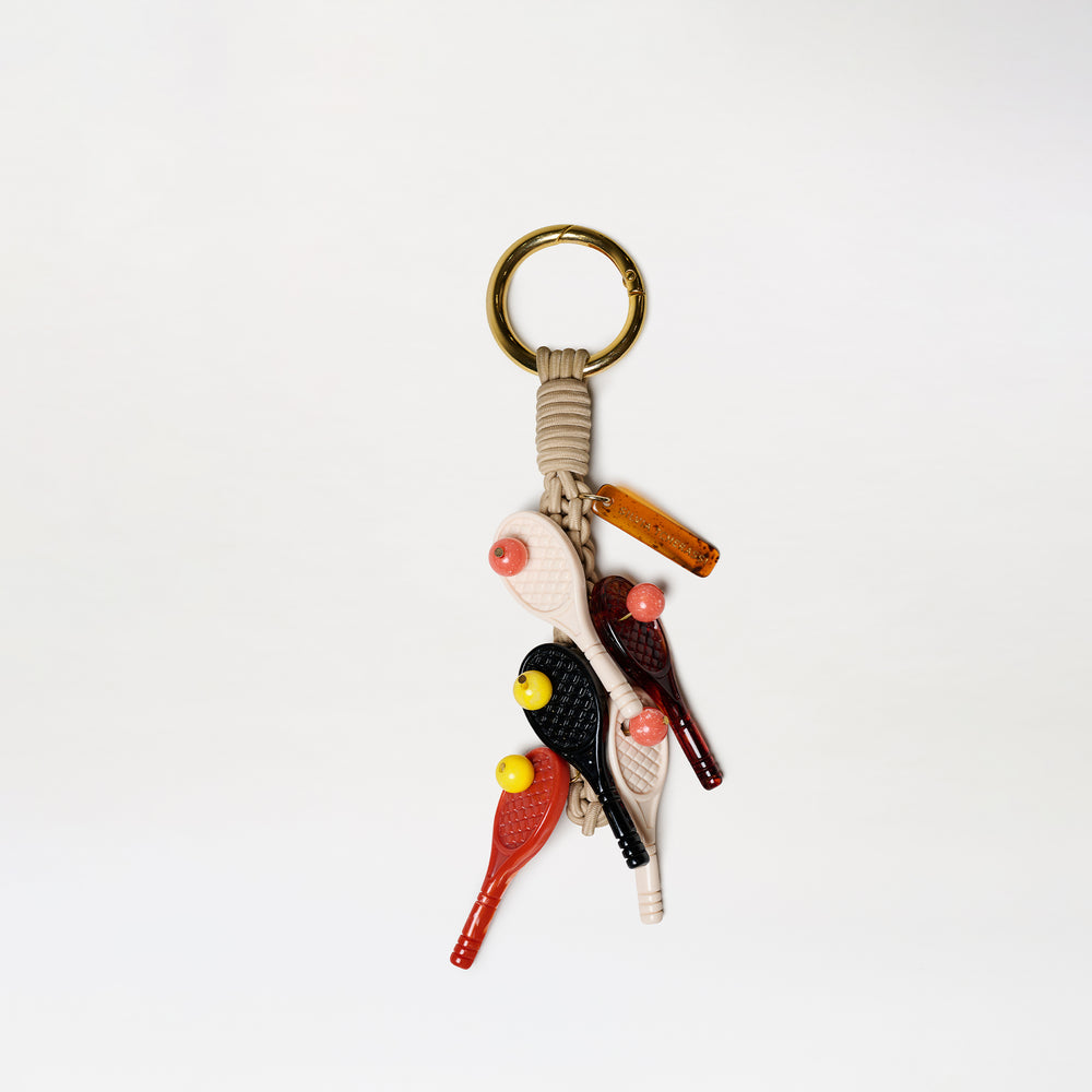 The Tournament Charm keychain features a gold ring, beige cord, and five bird-shaped charms in black, red, and beige with yellow beaks. A small brown rectangular tag is attached. All items are displayed against a white background.