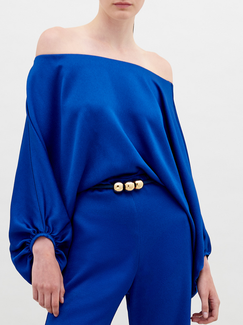 A person stands against a plain background wearing an off-the-shoulder blue top with wide sleeves and matching wide-leg pants, accessorized with a belt that has two large gold embellishments. Pre-order the Theresa Jumpsuit Royal Blue now to ensure you receive your outfit by the ship by date of November 15th, 2024.