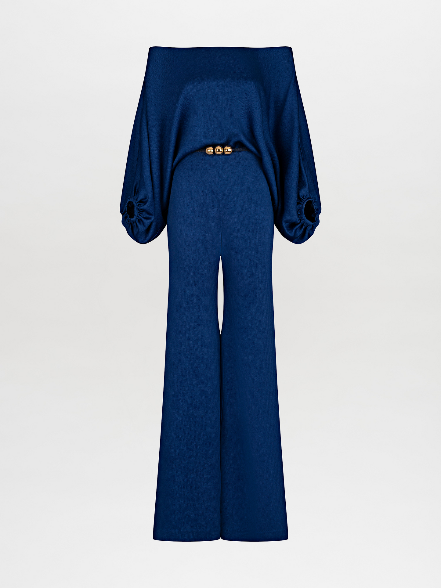 A person stands against a plain background wearing an off-the-shoulder blue top with wide sleeves and matching wide-leg pants, accessorized with a belt that has two large gold embellishments. Pre-order the Theresa Jumpsuit Royal Blue now to ensure you receive your outfit by the ship by date of November 15th, 2024.