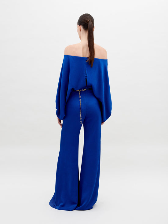 A person stands against a plain background wearing an off-the-shoulder blue top with wide sleeves and matching wide-leg pants, accessorized with a belt that has two large gold embellishments. Pre-order the Theresa Jumpsuit Royal Blue now to ensure you receive your outfit by the ship by date of November 15th, 2024.