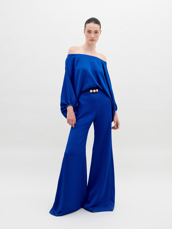 A person stands against a plain background wearing an off-the-shoulder blue top with wide sleeves and matching wide-leg pants, accessorized with a belt that has two large gold embellishments. Pre-order the Theresa Jumpsuit Royal Blue now to ensure you receive your outfit by the ship by date of November 15th, 2024.