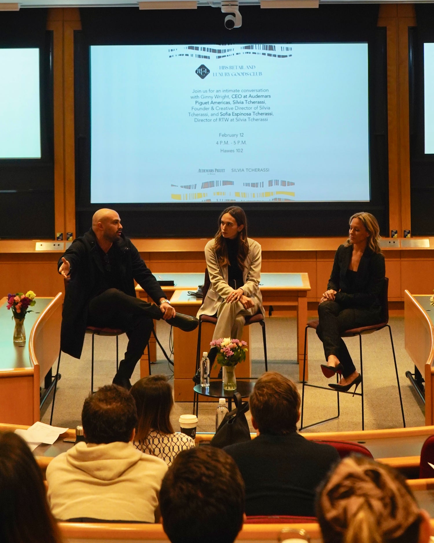 Three people sitting on stage in front of an audience at a lecture hall. A presentation screen is visible in the background.