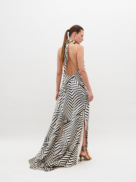 A person stands wearing a Tila Dress Beige Black Stripes with a high neckline, accessorized with a large floral detail at the shoulder, and gold footwear. This stunning outfit is available for pre-order and will ship by November 15th, 2024.