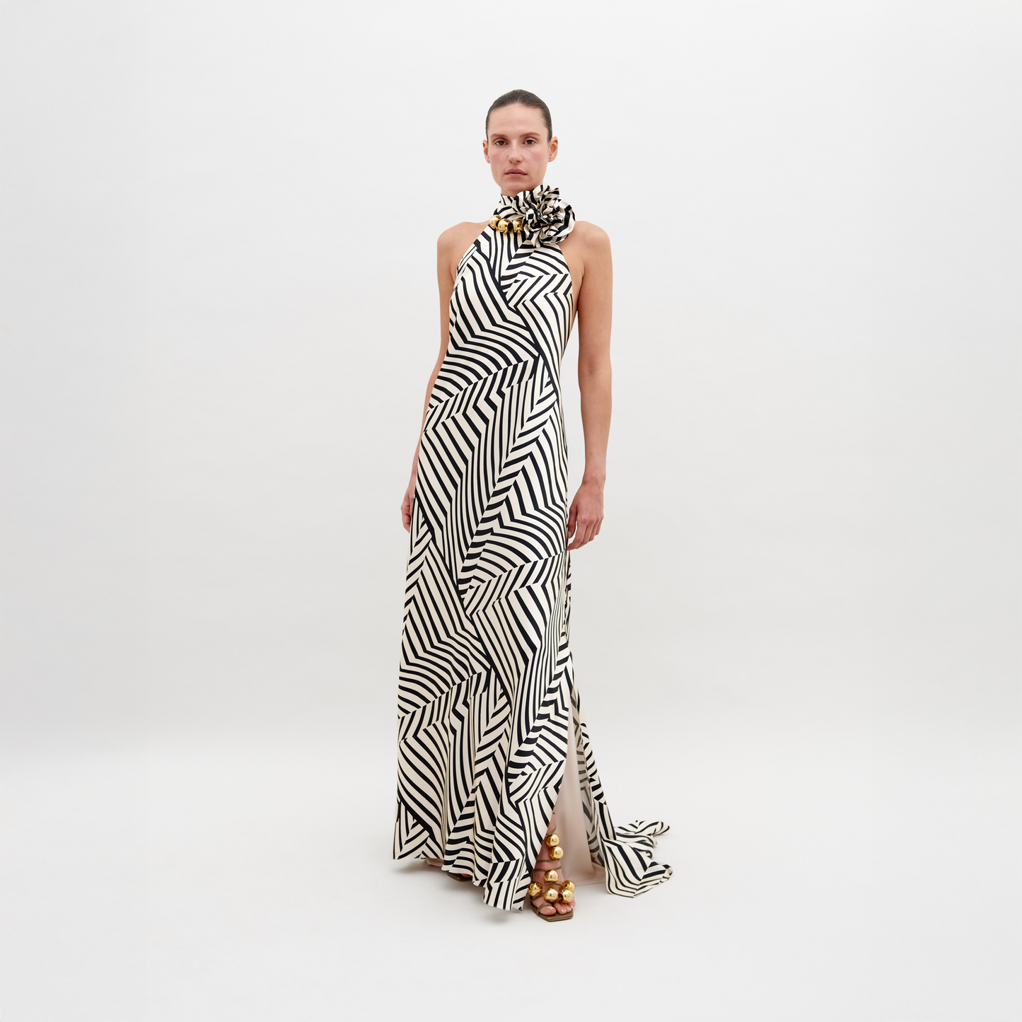 A person stands wearing a Tila Dress Beige Black Stripes with a high neckline, accessorized with a large floral detail at the shoulder, and gold footwear. This stunning outfit is available for pre-order and will ship by November 15th, 2024.