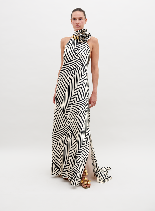 A person stands wearing a Tila Dress Beige Black Stripes with a high neckline, accessorized with a large floral detail at the shoulder, and gold footwear. This stunning outfit is available for pre-order and will ship by November 15th, 2024.