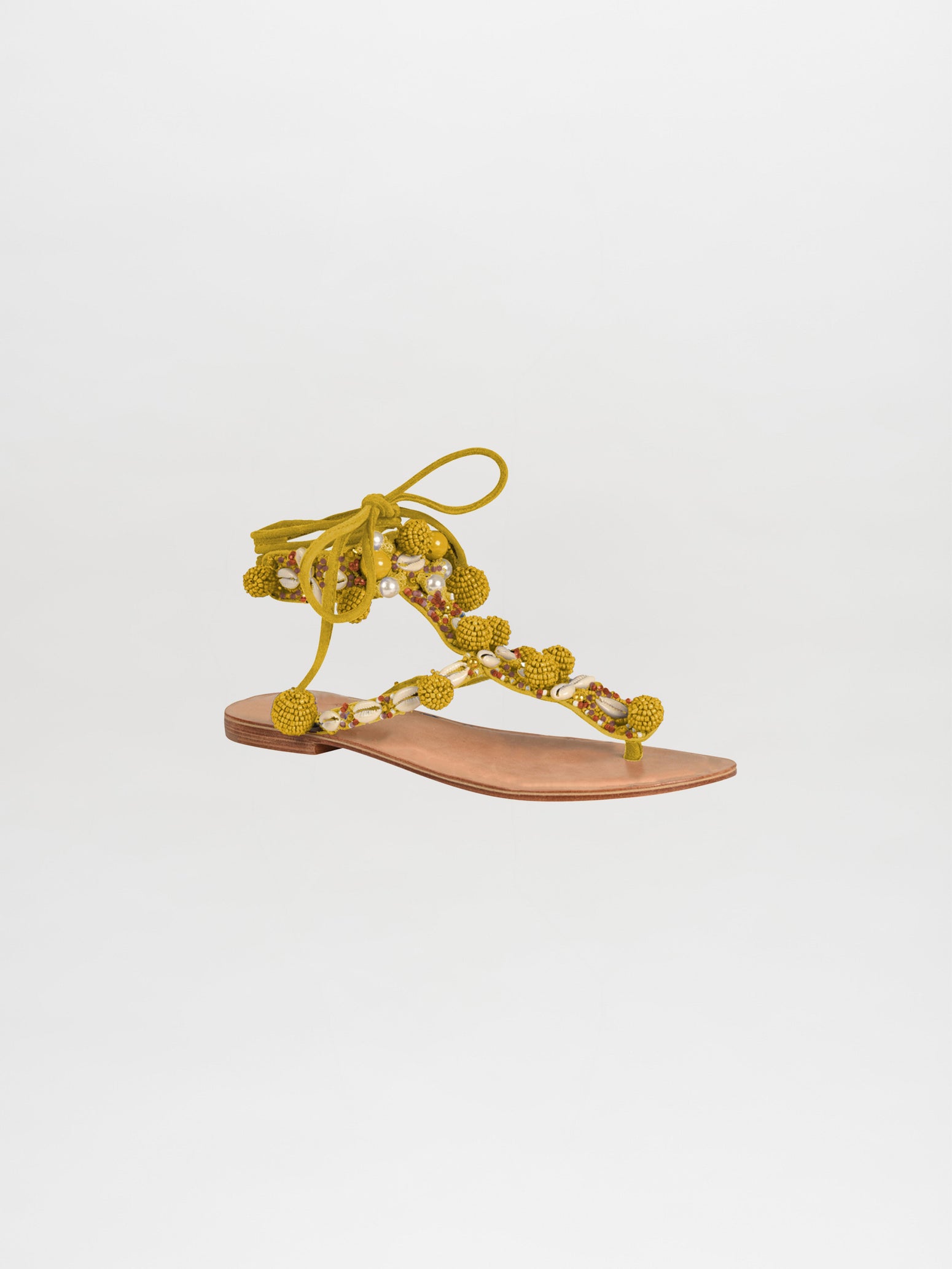 The Tisha Sandals Mustard feature a flat sandal with a brown leather sole, adorned with yellow pom-poms and beads, featuring olive green straps that tie around the ankle with added beading and seashell details.