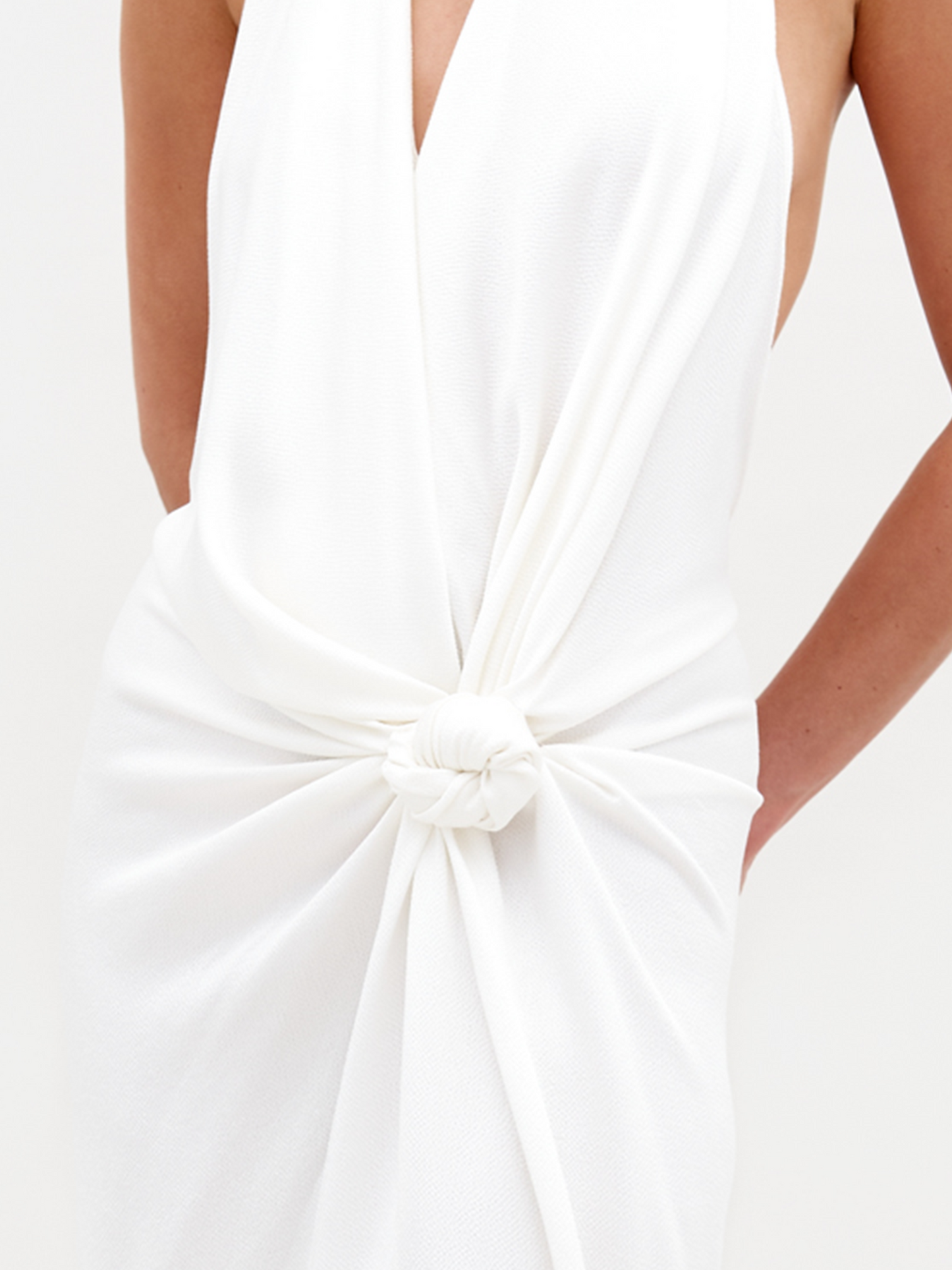 Person standing against a white background, wearing the Torgiano Dress White, which features a sleeveless halter design with a knot detail at the waist and a front slit. Pre-order now to ensure your dress is shipped by February 15th, 2025.