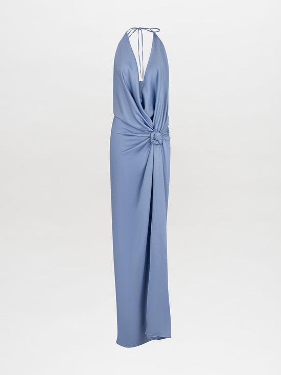 A person in the Torgiano Dress Periwinkle, a stunning blue halter-neck design with a central knot and an elongated hem, stands against a plain white background. This elegant piece is available for pre-order now and will ship by February 15th, 2025.