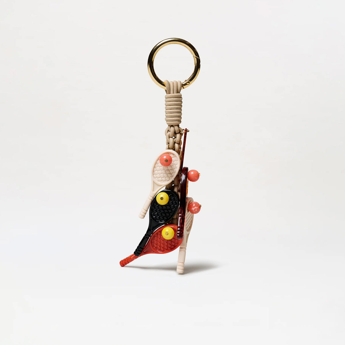 The Tournament Charm is a decorative keychain featuring a gold-tone ring, beige braided cord, and miniature red and black tennis racket charms, as well as small balls and sticks, all arranged in a playful and sporty design on a white background.