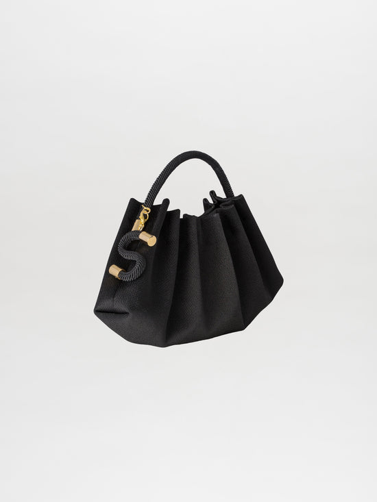 The Trapezius Clutch Black, a chic pleated handbag made from luxurious crepe fabric, showcases a curved handle and gold accents, elegantly displayed against a white background.