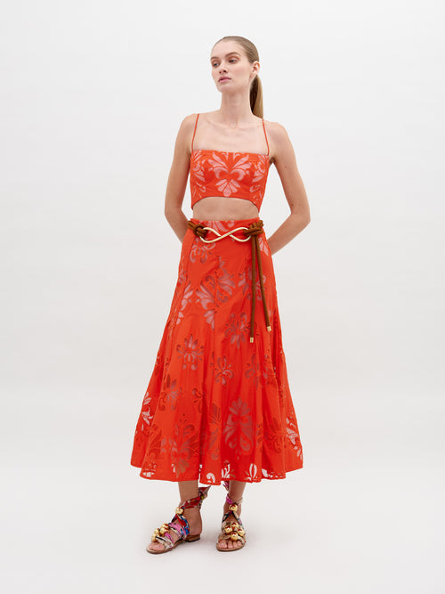 A woman stands wearing a Grettel Skirt Rouge Embroidery with a bright orange floral-patterned two-piece outfit and colorful sandals. She poses against a plain white background, available for pre-order starting November 15th for the 2024 collection.