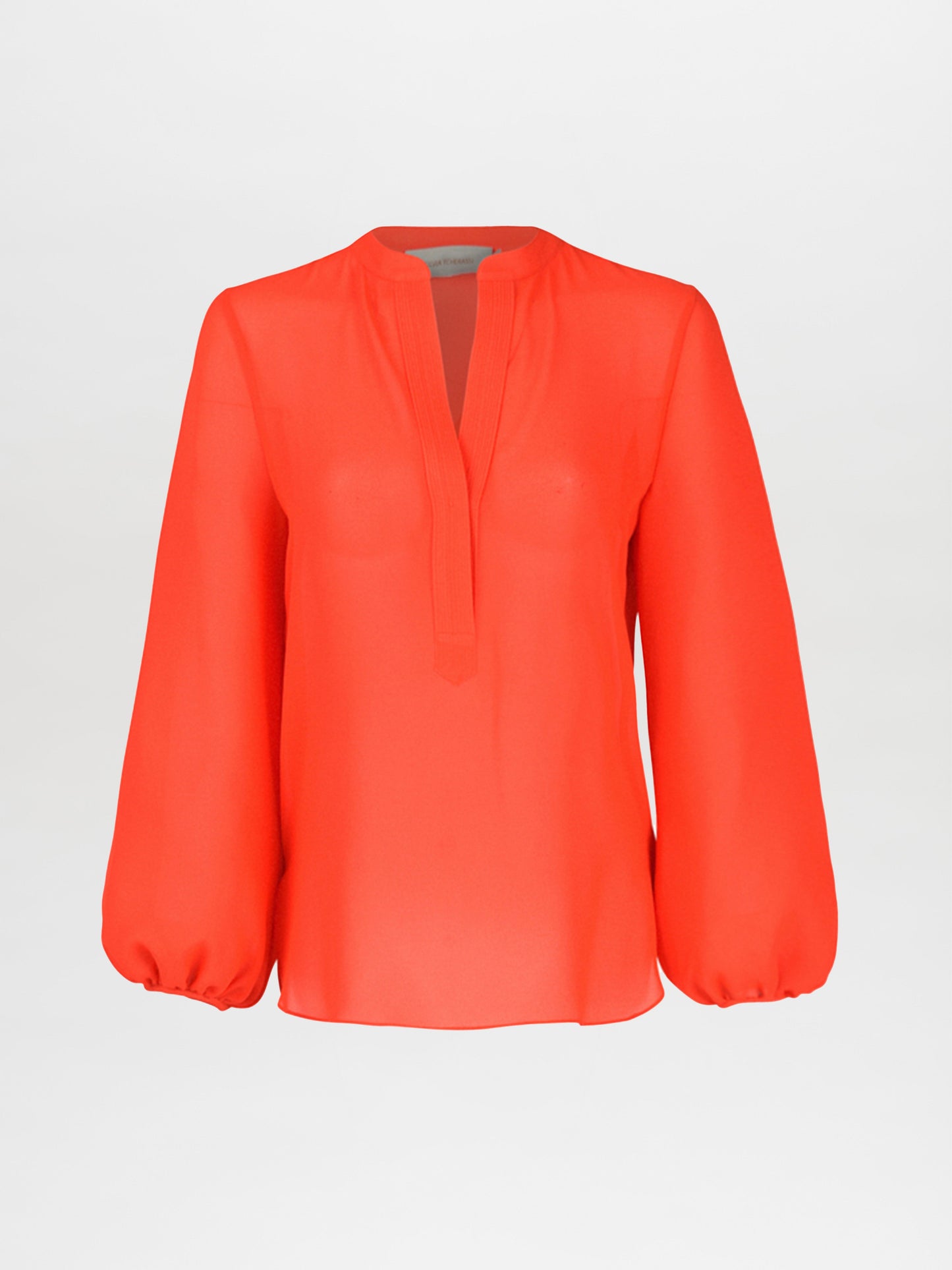 A person wearing a Triora Blouse in tangerine and a colorful asymmetrical skirt with layered draping stands against a neutral background.