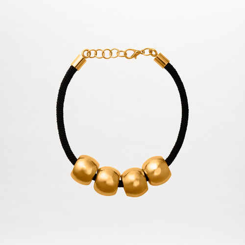 The Tristana Necklace is a black 3D cord choker featuring four large round gold beads with intricate details and a secure gold lobster clasp, all beautifully crafted against a plain backdrop. Made in Colombia.