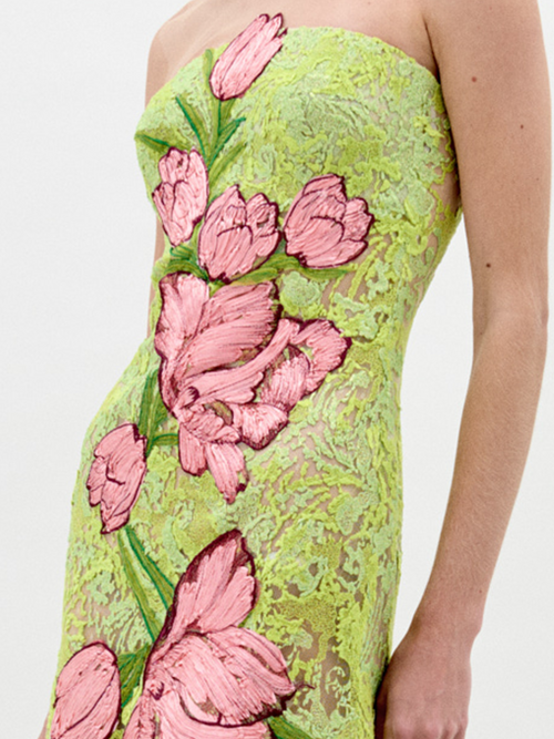 A woman stands wearing the Tulip Dress Rose Tulips, a strapless, green lace dress adorned with large pink floral designs, looking to her left against a plain light background. Pre-order now for exclusive access; shipping by February 1st, 2025.