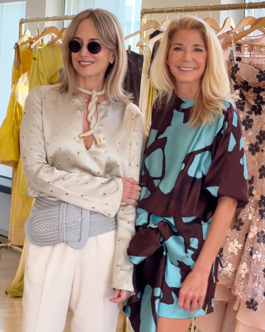 Two women standing in a clothing store, surrounded by colorful dresses on hangers. One wears sunglasses and light clothing; the other wears a patterned dress. Both are smiling.