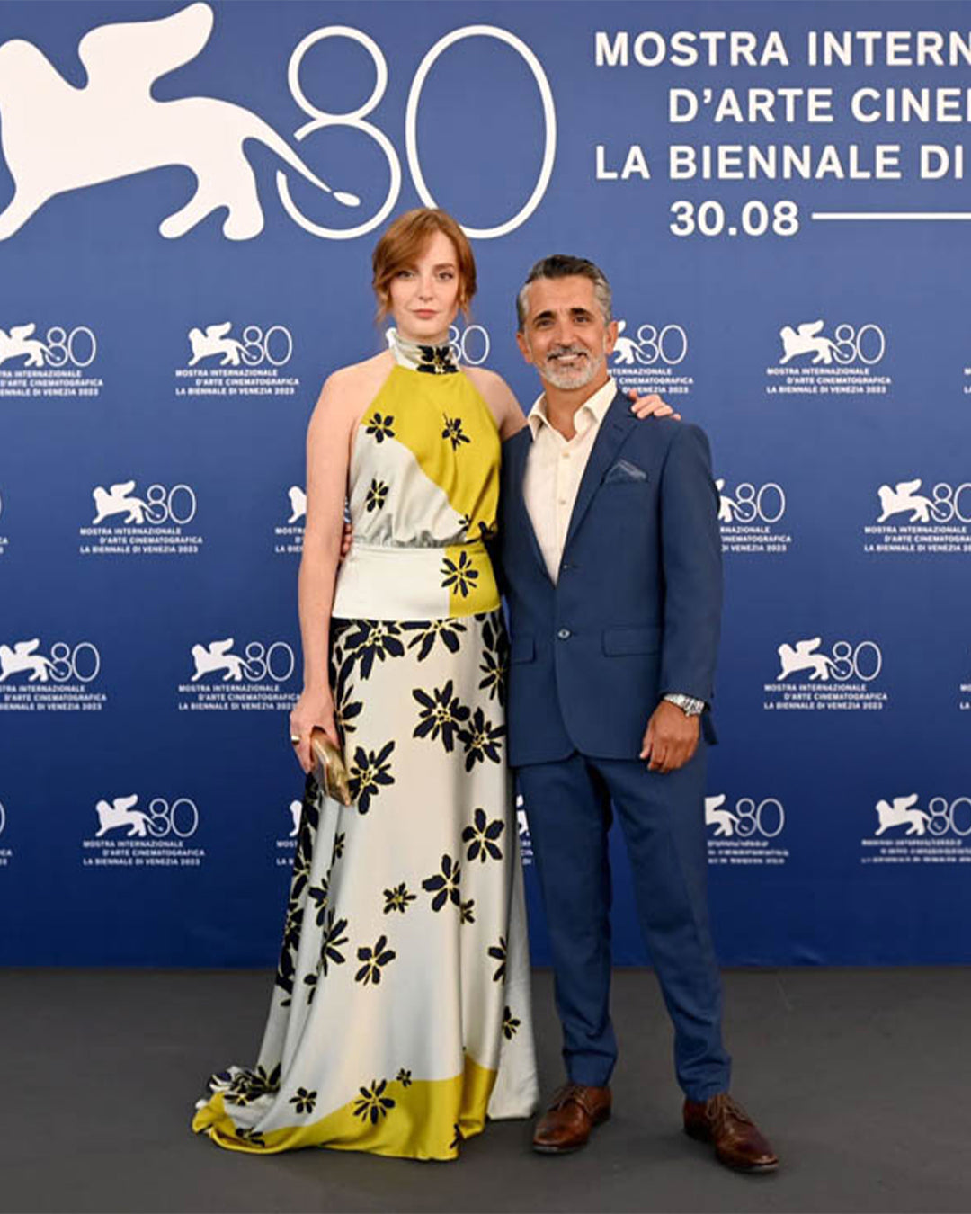 Two individuals, one in a floral dress and the other in a blue suit, stand in front of a "Mostra Internazionale d'Arte Cinematografica" backdrop.