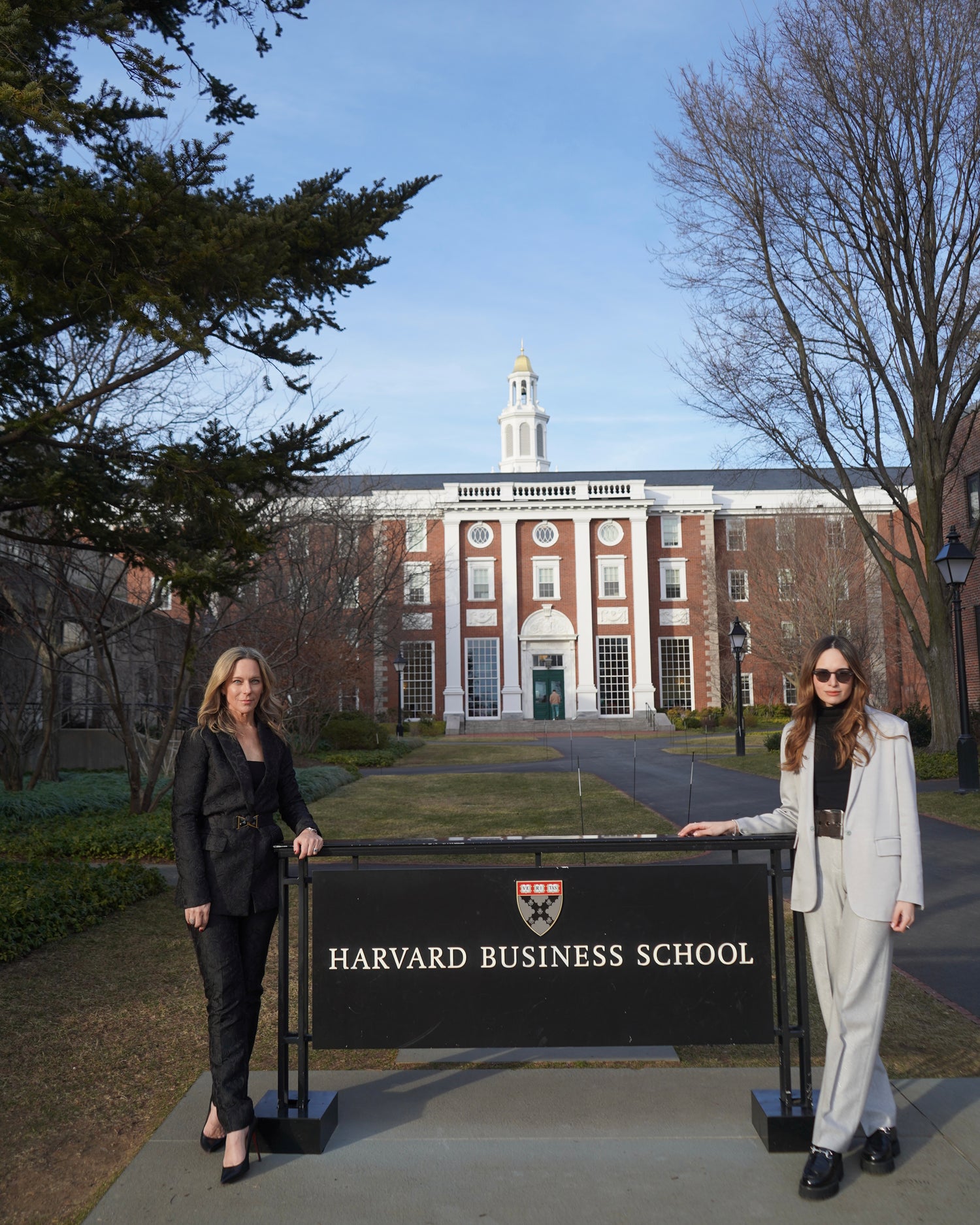 Two people stand in front of a Harvard Business School sign, with a campus building and trees in the background.