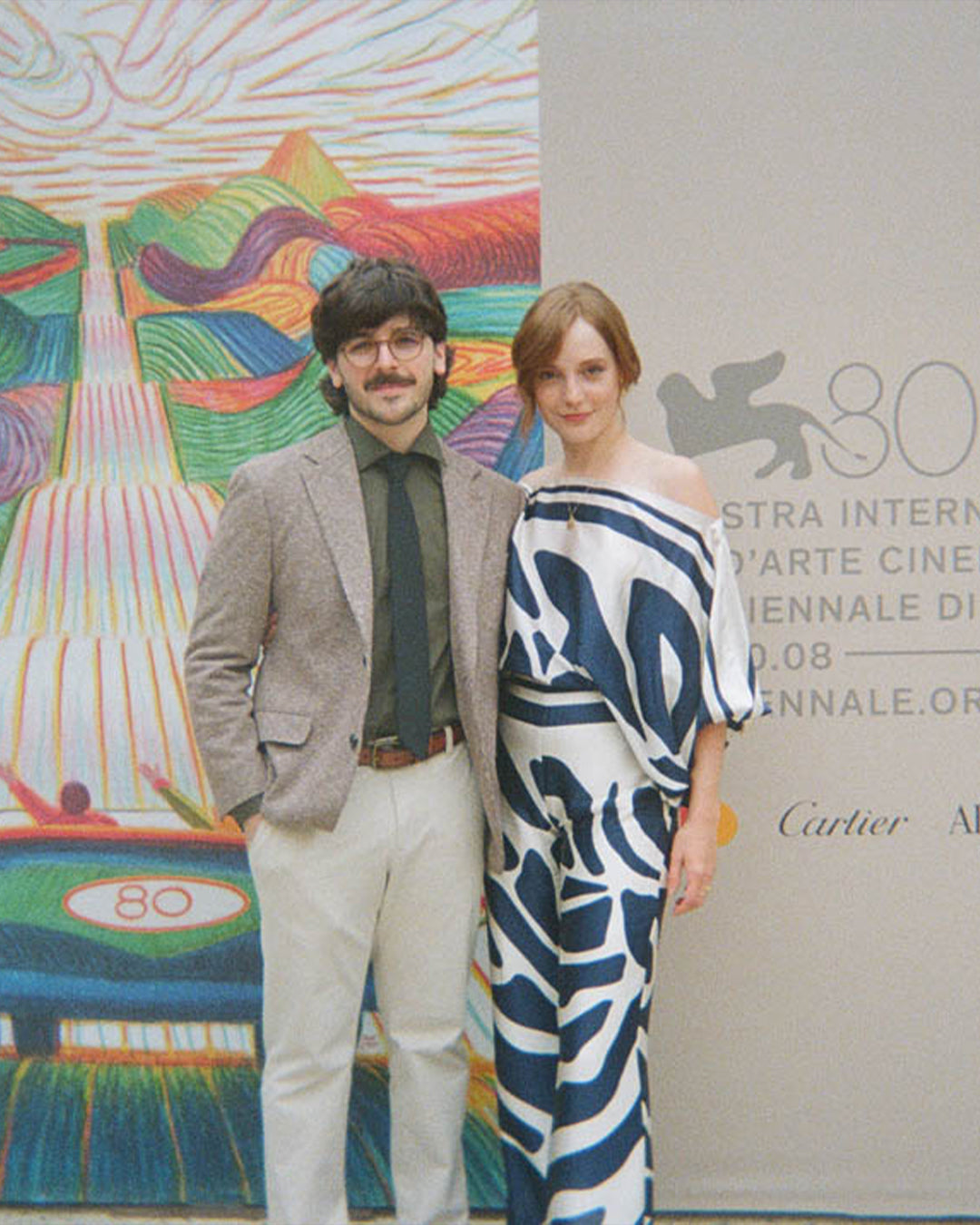 Two people stand in front of a colorful artistic backdrop and a sign that reads "80", dressed in formal attire.