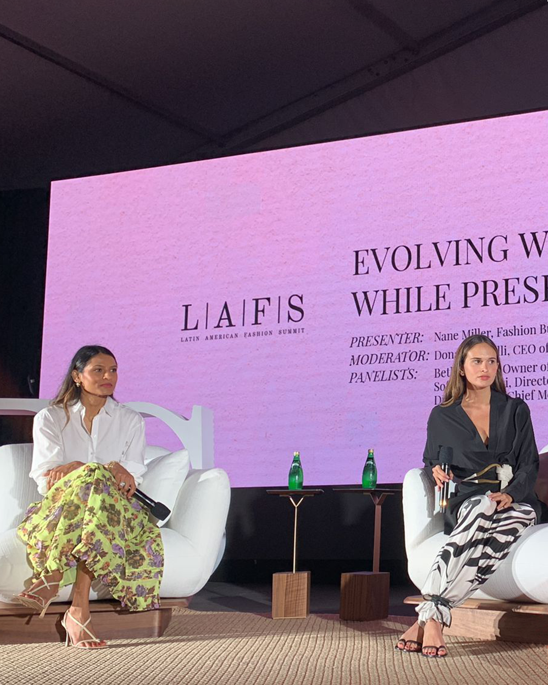 Two women seated on a stage with a large pink screen behind them displaying text for the Latin American Fashion Summit. Bottles of water are on a table between them.