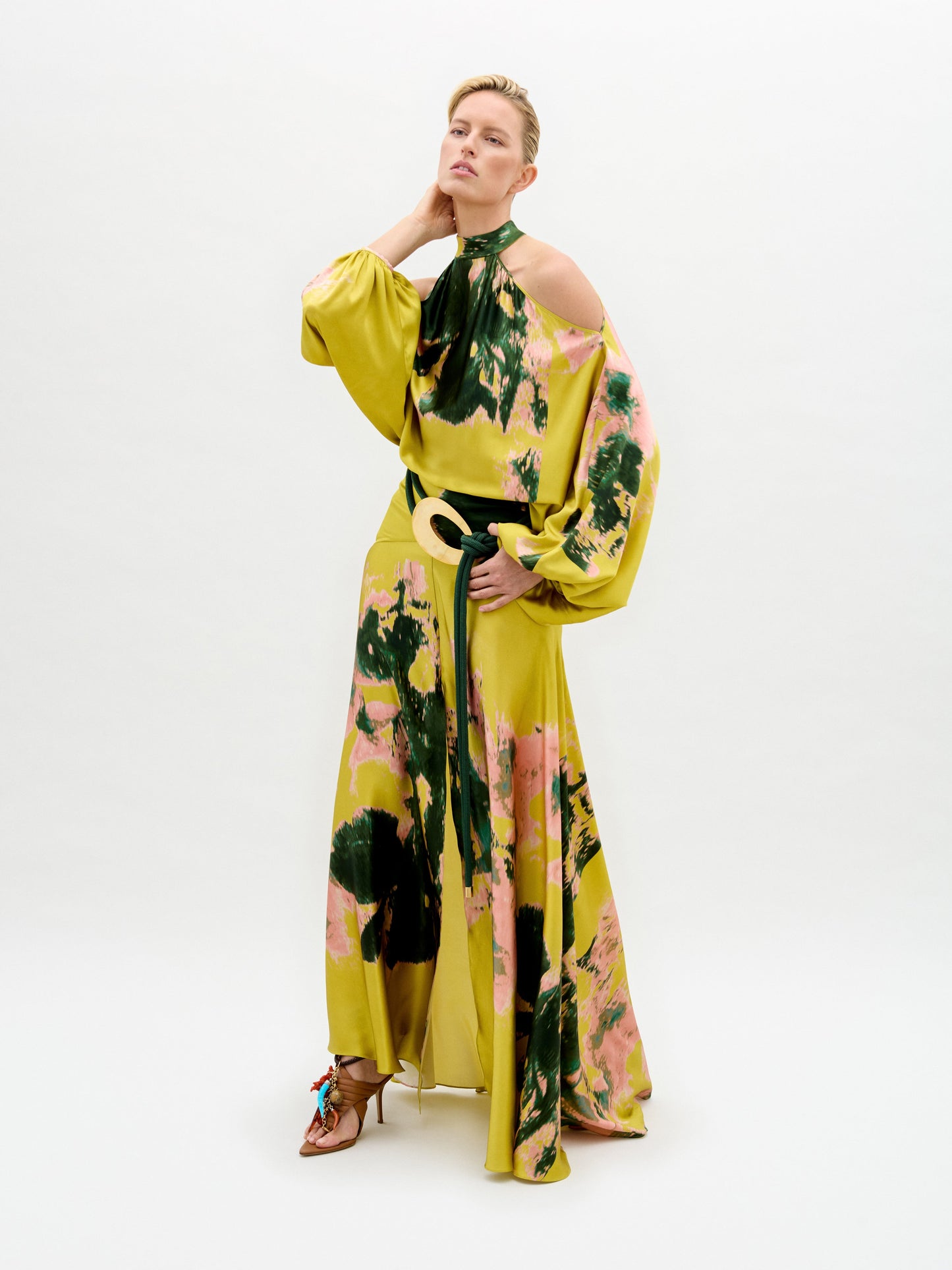 A person wearing the Valeria Dress, a long, colorful garment featuring a green and pink pattern, poses against a plain white background.