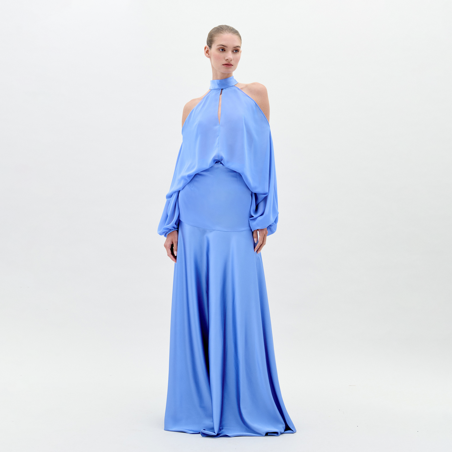 A person in a long, flowing Valeria Dress Sky Blue with shoulder cutouts and a high neckline stands against a plain white background. This elegant attire is available for pre-order now and will ship by February 1st, 2025.