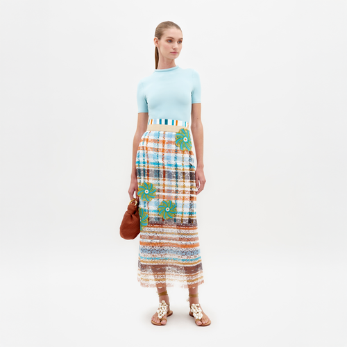 A woman stands against a plain background wearing a light blue top, the Shadia Skirt Multi Color Stripes, and white sandals. She holds a small brown bag, ready for her February 15th event in 2025.