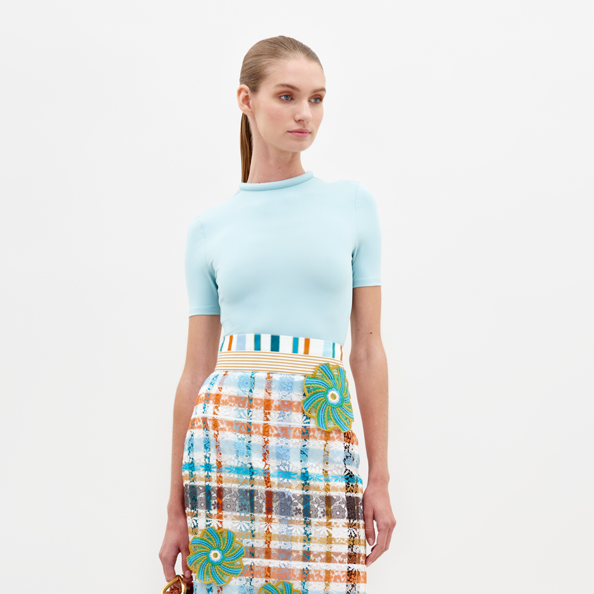 A woman dressed in a fitted Varinia Tshirt Turquoise and a multicolored plaid skirt stands against a plain white background, holding a small brown item in her right hand.